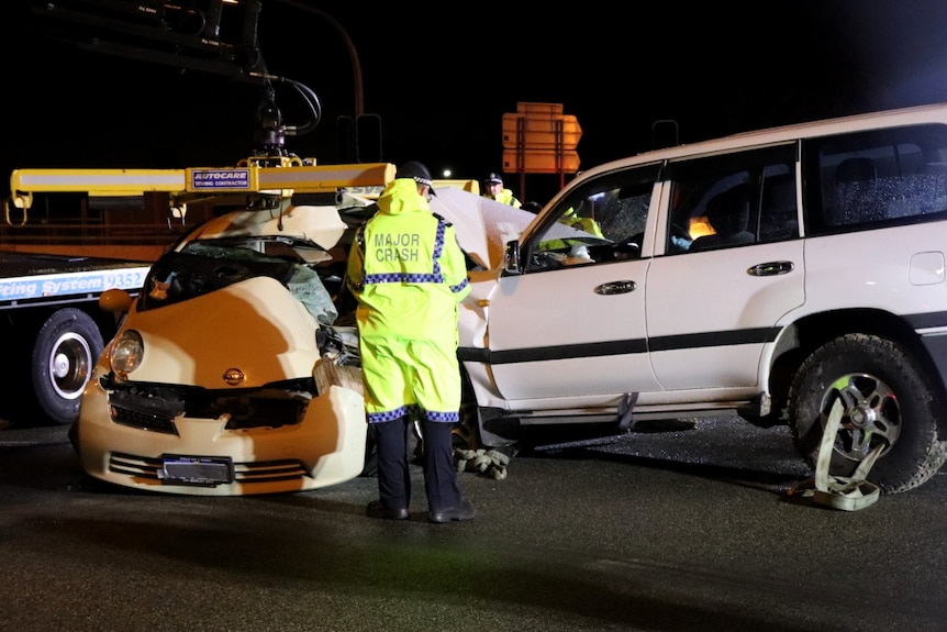 A police officer stands in front of a yellow hatchback which has been t-boned by a white four-wheel drive.