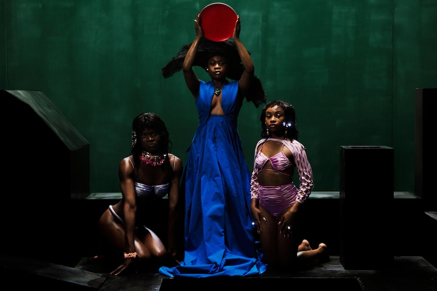 a group of three women with one in the centre hold what looks like a drum as the other two kneel beside her