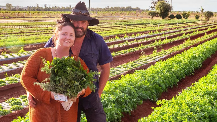 A woman and man stand in a field of herbs holding a bunch of herbs.