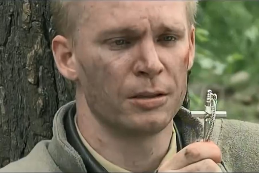 A blonde, fair-skinned man with a dirty face looks at a cross necklace with a look of despair