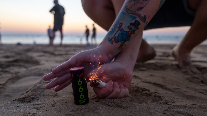 A man strikes a lighter and places it next to a firework on the beach.