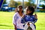 A little girl in a purple floral dress is pushed on a swing with her mother in the foreground slightly out of focus