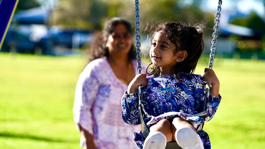 A little girl in a purple floral dress is pushed on a swing with her mother in the foreground slightly out of focus