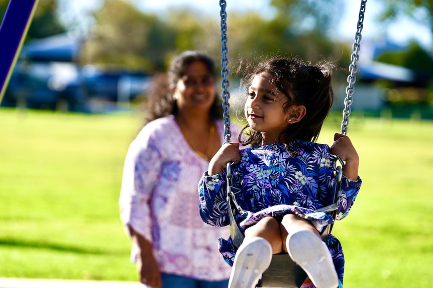 A little girl in a purple floral dress is pushed on a swing with her mother in the foreground slightly out of focus