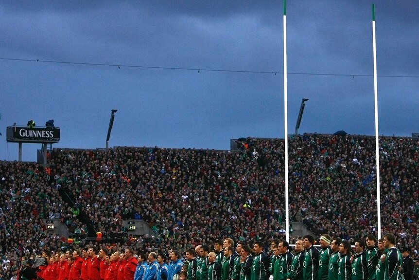 Players wearing red and green tracksuits line up in front of fans on a terrace with rugby posts in the background