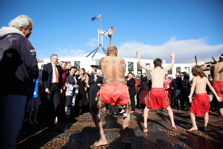 Albanese and a crowd of others watch several Indigenous men in traditional clothing dance outside Parliament.