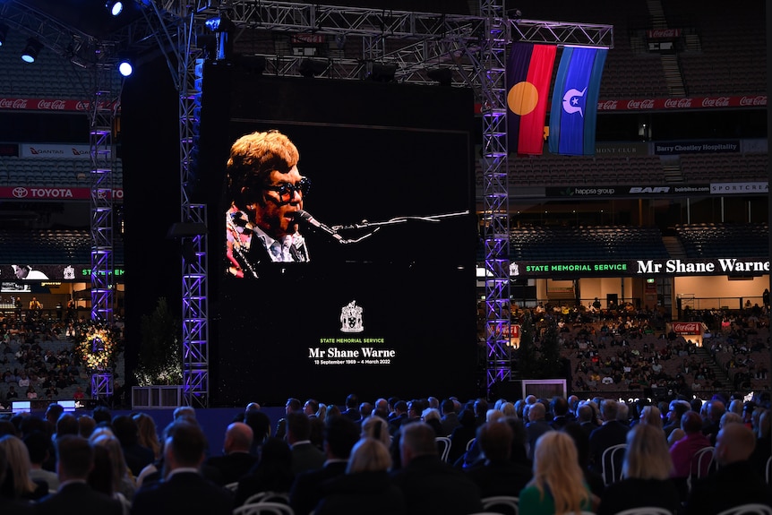 A picture of Sir Elton John on a screen with a crowd in the foreground.