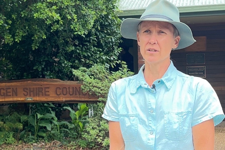 Councillor Jennie Fenton wearing a short sleeve shirt and a brimmed hat standing in front of a sign for Bellingen Shire Council.