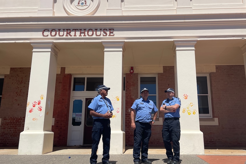 three police officers in uniform stand oustide a courthouse in the daytime where red and yellow handprints have been painted