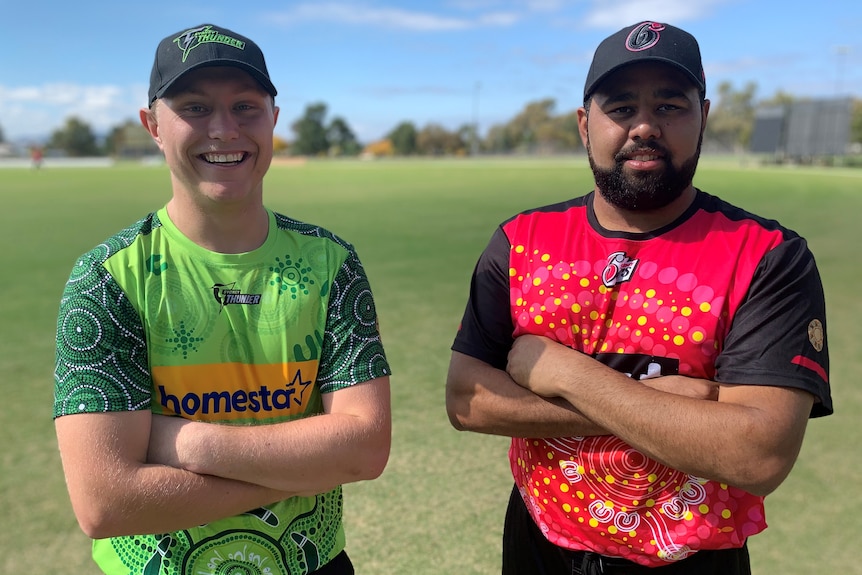 Two cricketers in colourful uniforms stand side by side, grinning.