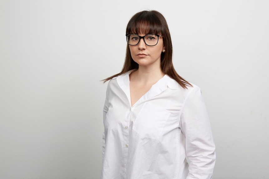 A brunette woman standing up wearing a white business shirt and wearing black glasses
