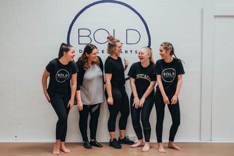 Five young women smile as they stand in a line in a dance studio.