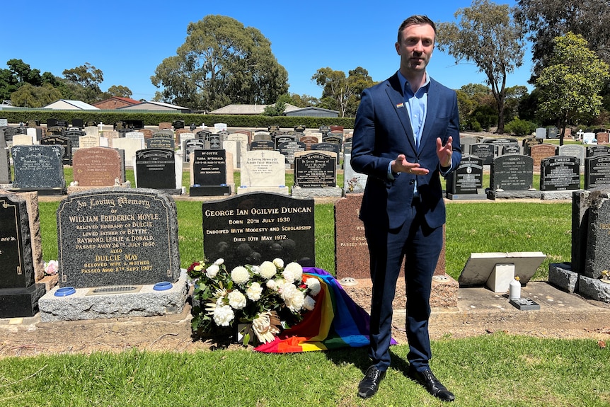 A man wearing a suit without a tie standing in a cemetery next to a grave with flowers on it