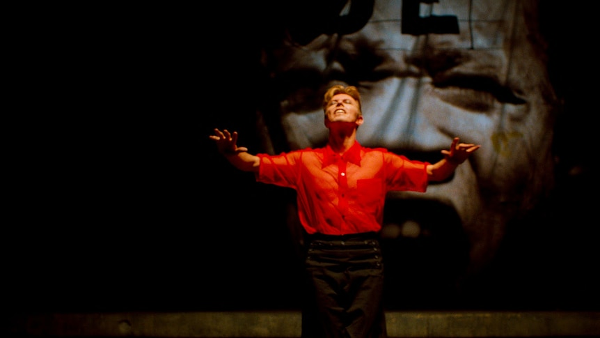 David Bowie wearing a red shirt and black pants with short blonde hair stands on stage with arms lifted and head thrown back.