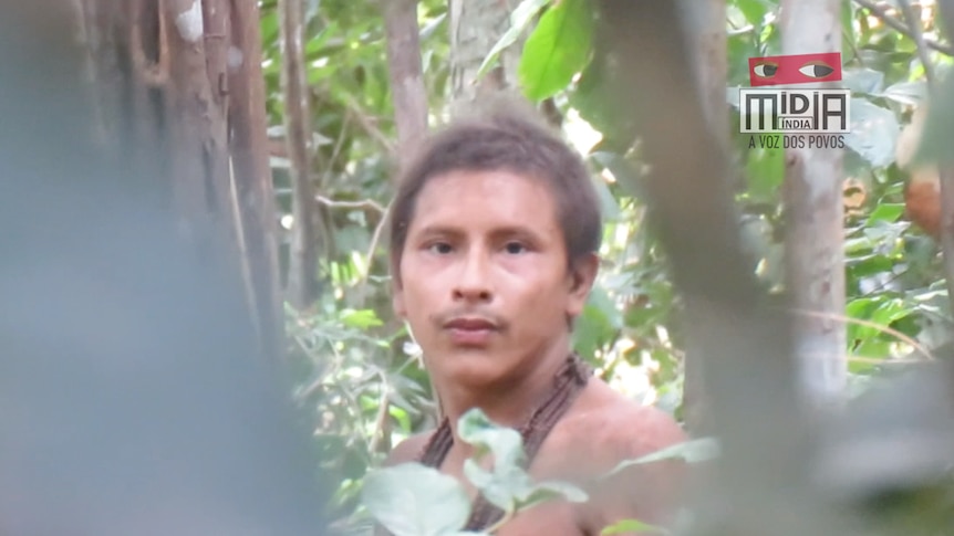 A man looks int he direction of a camera while stranding in the forest. There are leaves obscuring the view of the image.