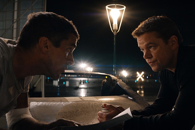 Christian Bale and Matt Damon face each other, learning forward on bonnet of vintage muscle car at night in garage.
