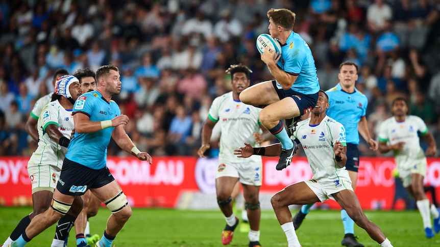 A rugby union player in a light blue jersey jumps in the air and catches the ball during a match