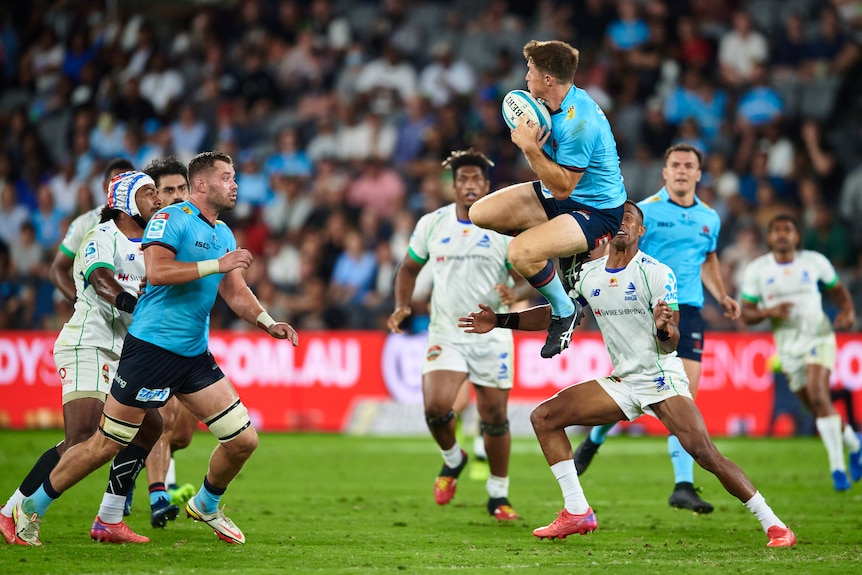 A rugby union player in a light blue jersey jumps in the air and catches the ball during a match