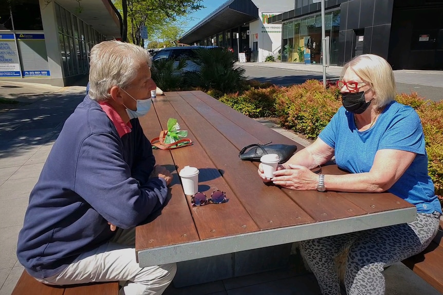 An older man and woman sit on a park bench with take away coffees talking.