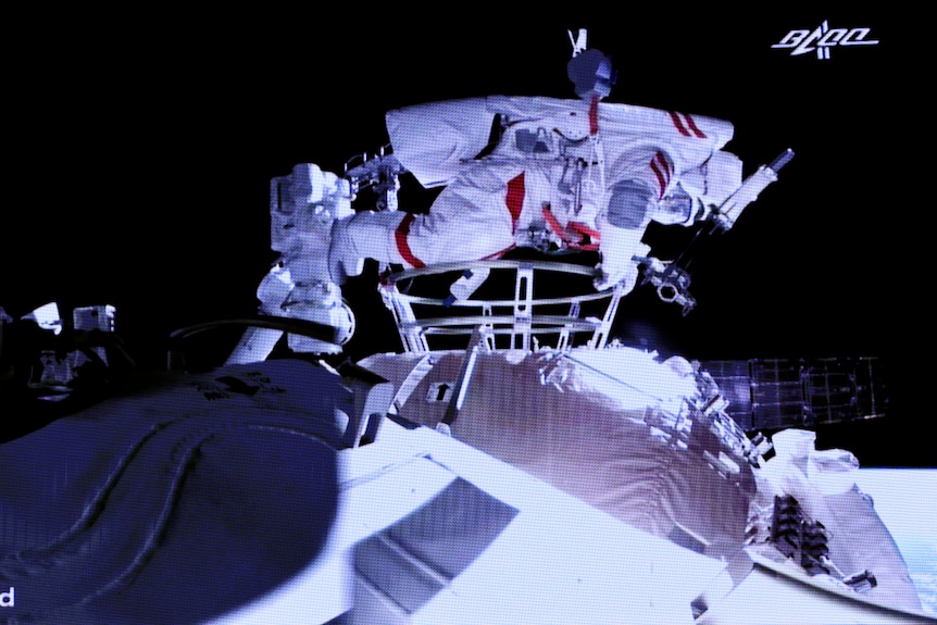 Una persona in una tuta spaziale squadrata aggrappata all'esterno di una stazione spaziale.