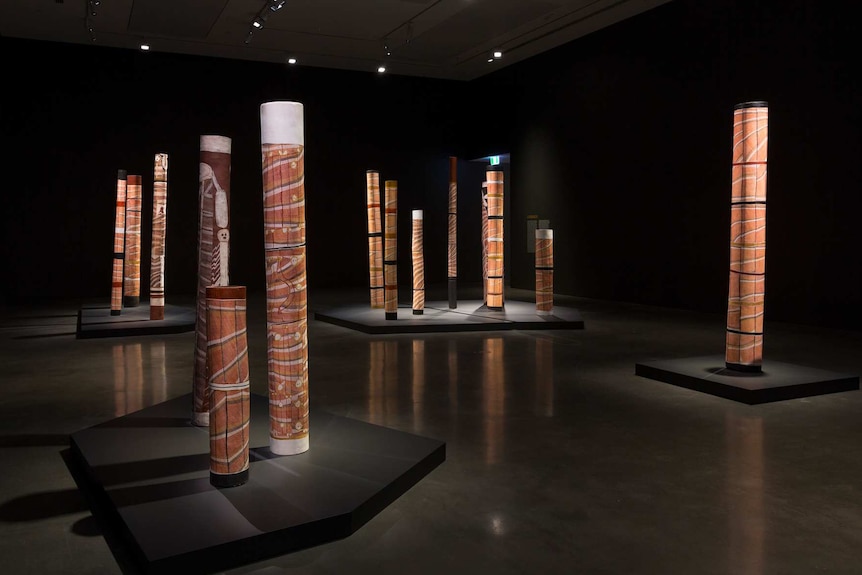 Darkened gallery featuring installation of upright painted "Lorrkon", Aboriginal burial logs.