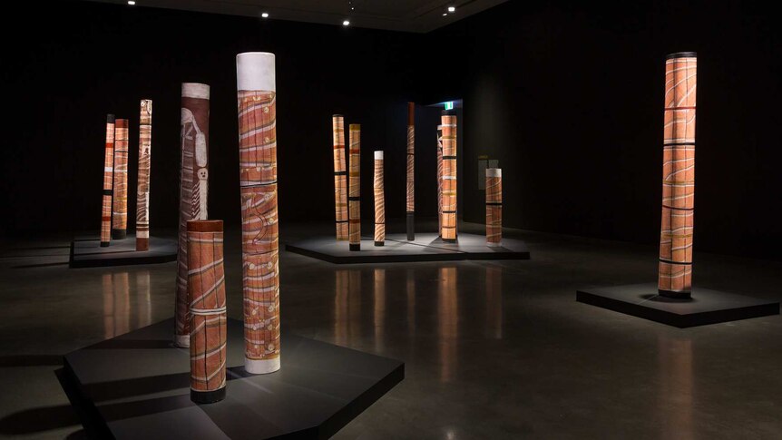 Darkened gallery featuring installation of upright painted "Lorrkon", Aboriginal burial logs.