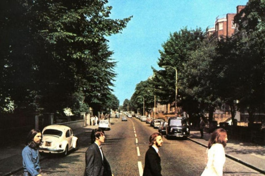 Four men in flared trousers, three with jackets, walk across a pedestrian crossing.