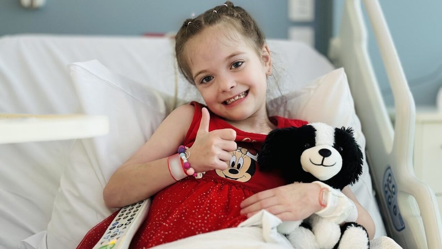 A young girl in a red dress lies in a hospital bed, smiling, giving the thumbs up signal, with a toy dog.  