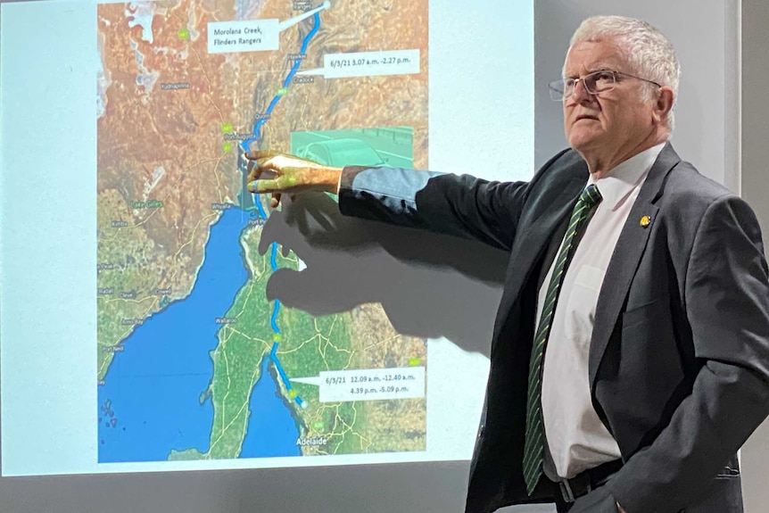 A man in a suit and with short white hair and spectacles points to a line on a map of South Australia.