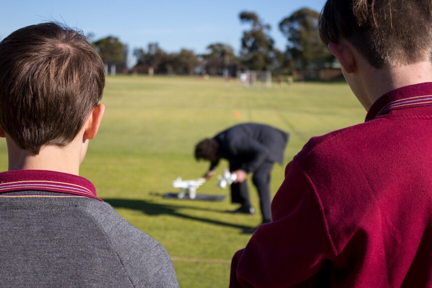 Looking between two students shoulders as instructor places drone on ground.