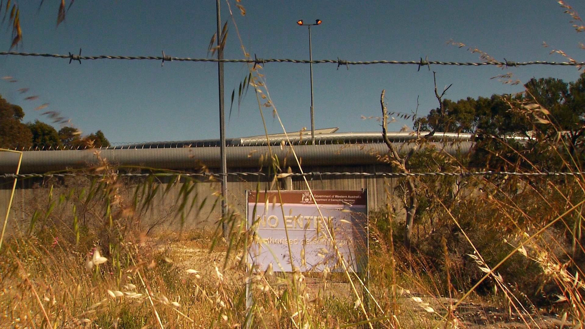 The walls of a youth detention centre through a barbed wire fence. A sign partially obscured by grass says 'no entry'.