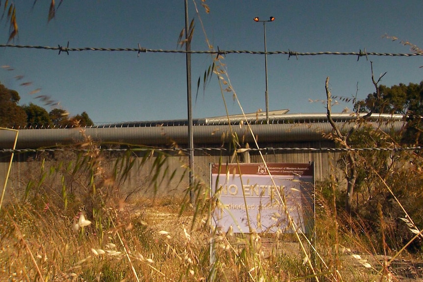 The walls of a youth detention centre through a barbed wire fence. A sign partially obscured by grass says 'no entry'.