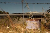 The walls of a youth detention centre through a barbed wire fence. A sign partially obscured by grass says 'no entry'.