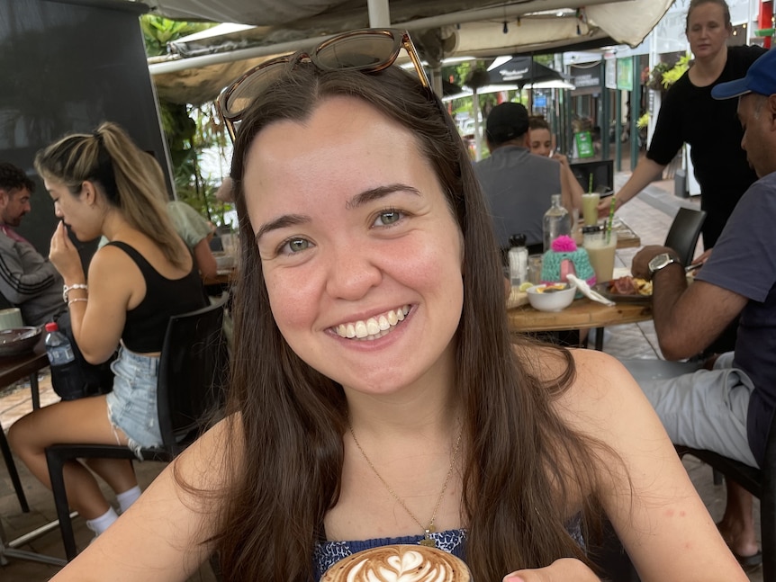 A smiling woman sits at a busy cafe holding a cup of coffee