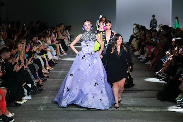 A woman in black walks at the head of a line of models in elaborate dress