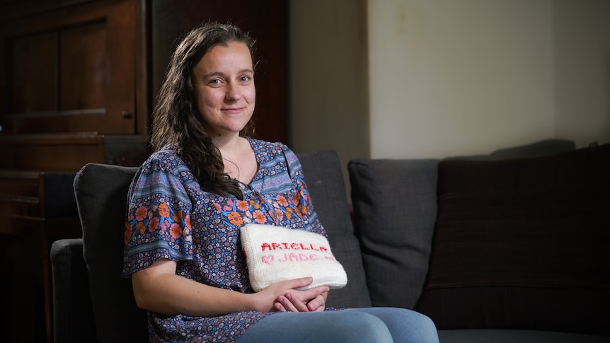 A woman with long brown hair sitting while holding a white cushion with the words ARIELLA JADE on it