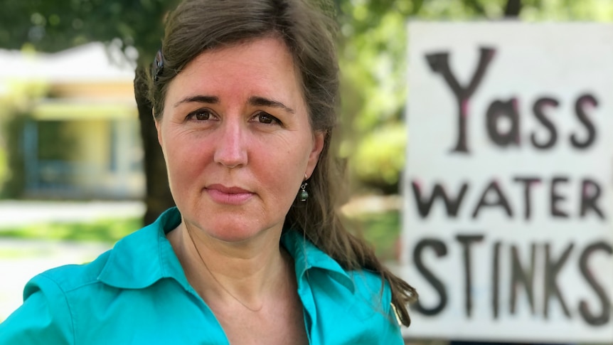 A woman has a blank stare standing in front of a sign that says "Yass water stinks"
