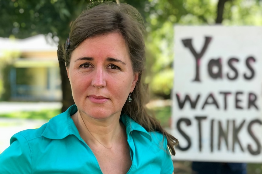 A woman has a blank stare standing in front of a sign that says "Yass water stinks"