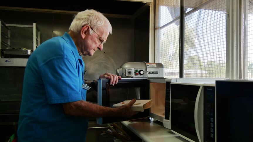 An grey-haired elderly man in a blue shirt puts a meal in a microwave over.