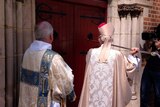 Archbishop Kay Goldsworthy, in dress robes, uses a staff to knock on door of St George's Cathedral as a church official watches.