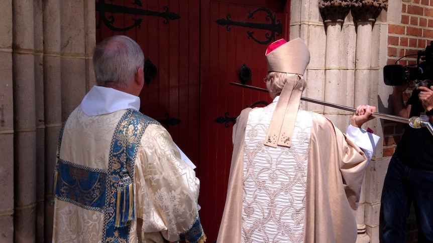 Archbishop Kay Goldsworthy, in dress robes, uses a staff to knock on door of St George's Cathedral as a church official watches.
