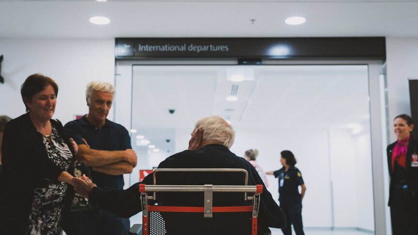 A man in a wheelchair photographed from behind, approaching a sliding door with an "international departures" sign above it.