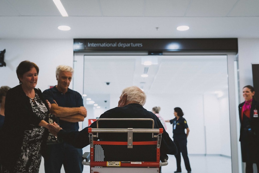 A man in a wheelchair photographed from behind, approaching a sliding door with an "international departures" sign above it.
