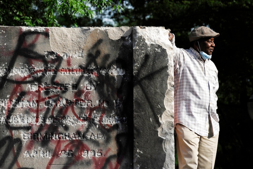 You look at a damaged base of a Confederate statue as an African American man leans on it and poses next to it.