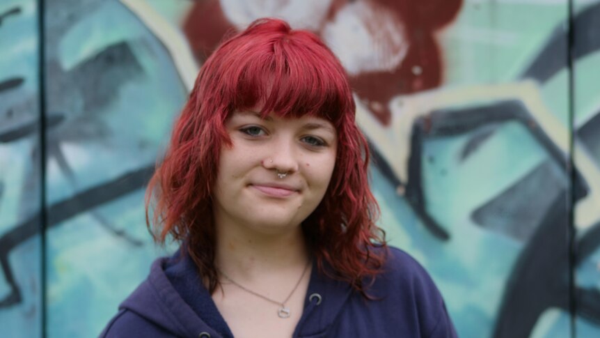 young girl with red medium length hair and short fringe, wearing a navy jumper, smiles with graffiti wall background