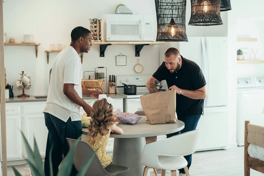 two men and a young girl at the kitchen table unpack groceries