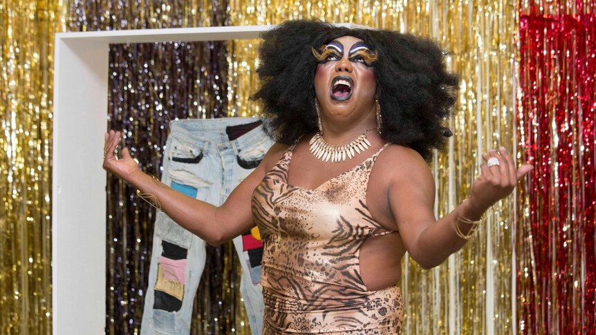 Drag queen standing in front of a tinsel curtain and jeans artwork
