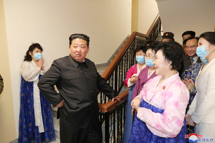 Kim Jong Un in a black suit with his hand on a railing as people gather around him inside a building