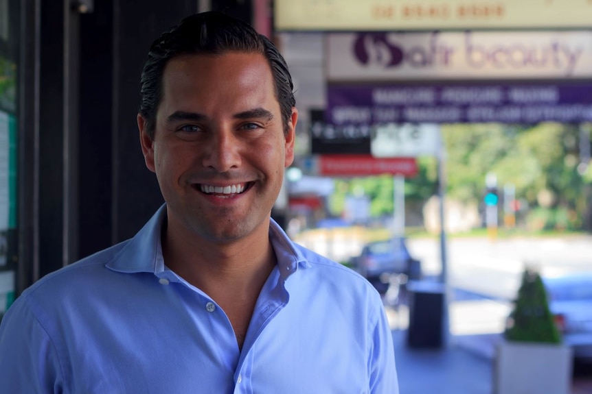 A smiling, dark haired man in a blue shirt standing on a city street.