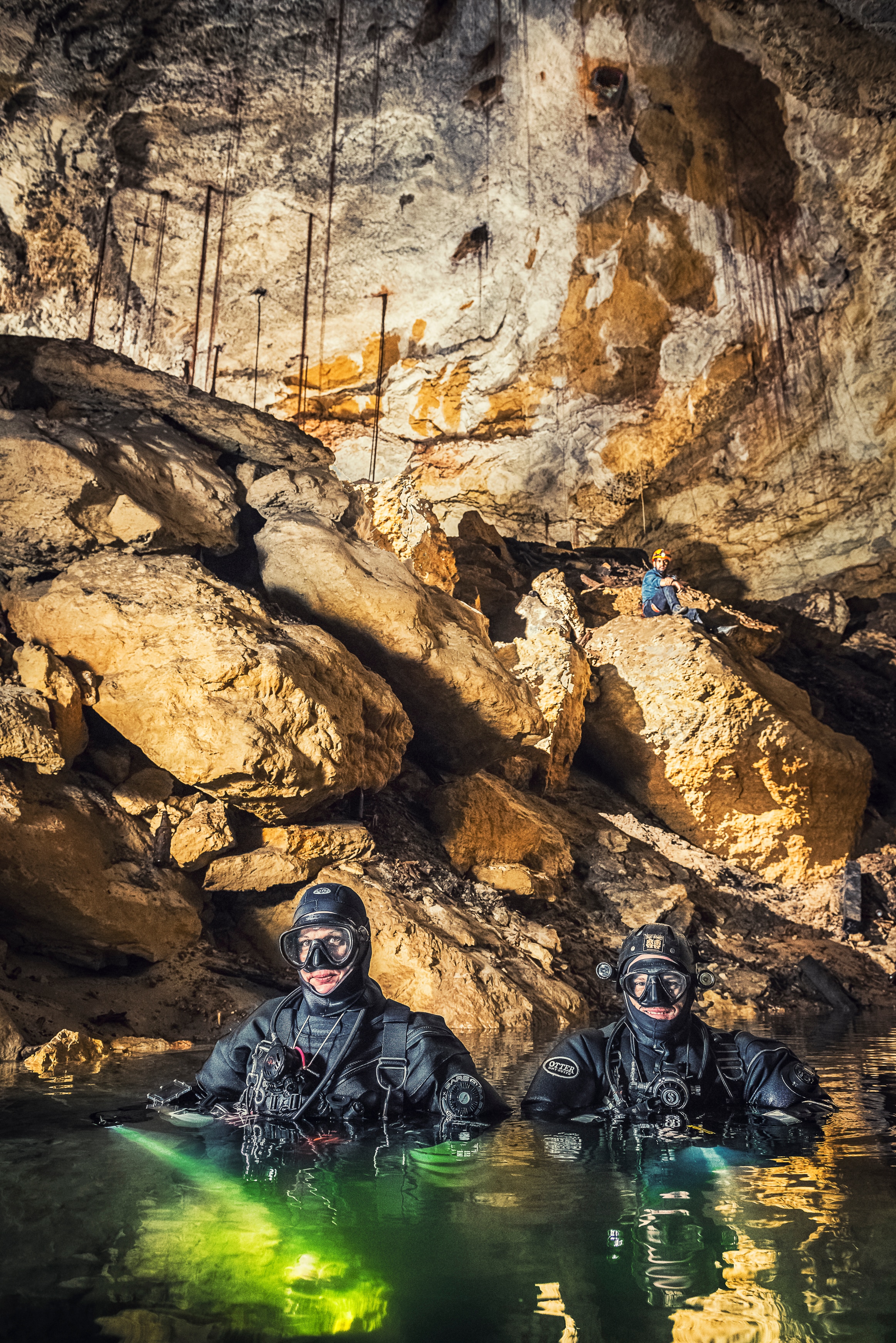 Two people wearing diving suits inside water inside a cave. 